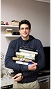 Smiling man holding a stack of books standing in an indoor setting with soft background.